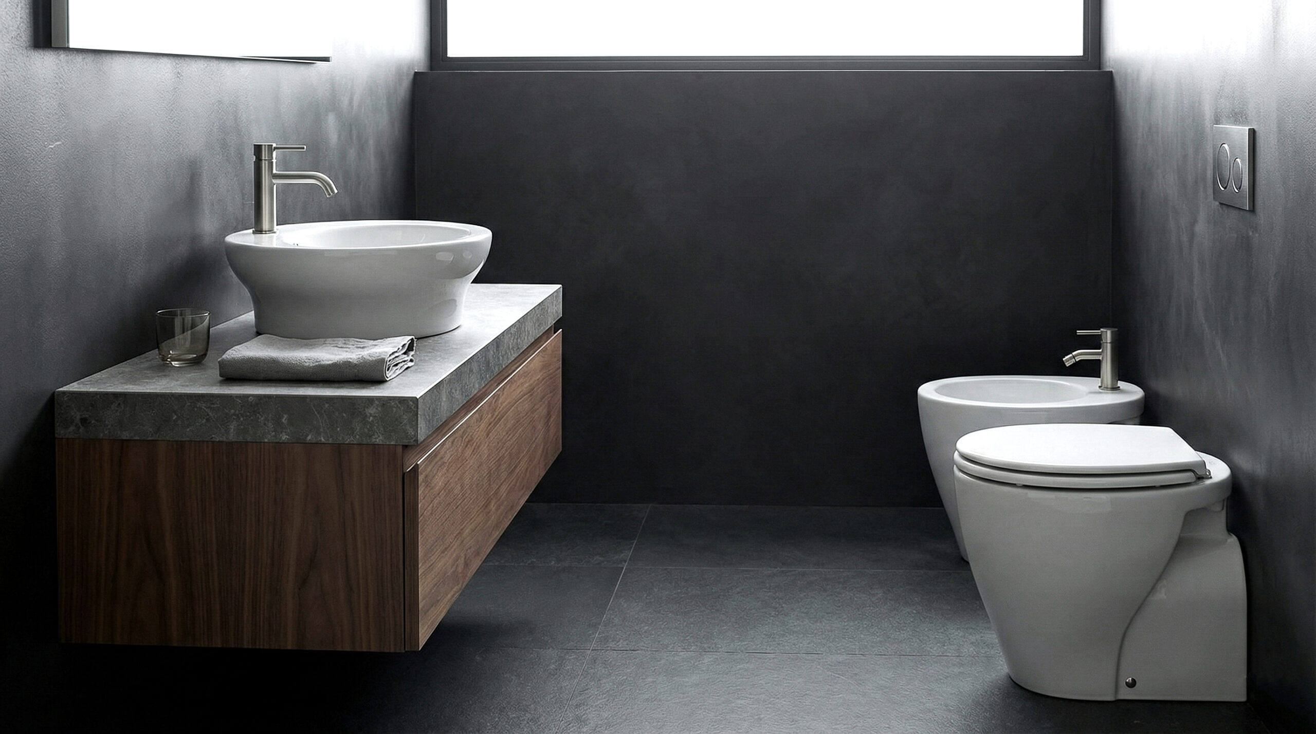 Lavabo d’appoggio su base sospesa con piano in pietra e sanitari coordinati in bagno moderno scuro dal design minimale.