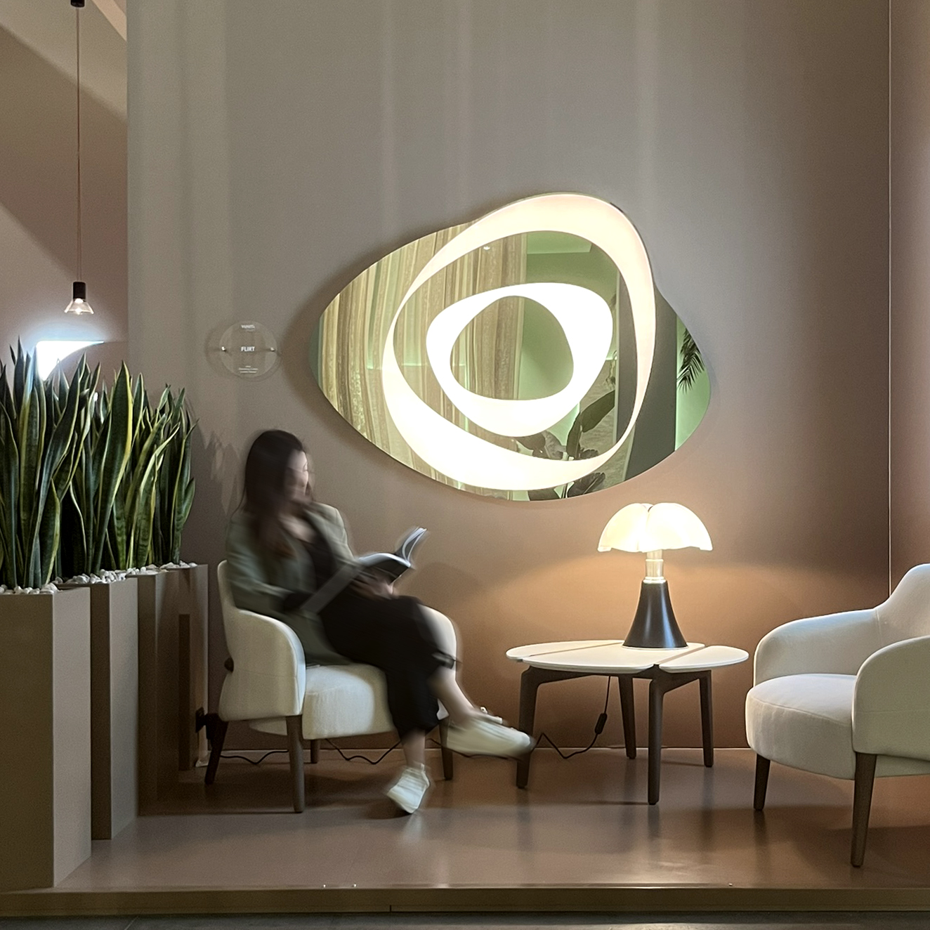Lounge con poltrone e tavolino, lampada da tavolo e grande specchio ovale con elementi luminosi concentrici su parete dai toni caldi.
