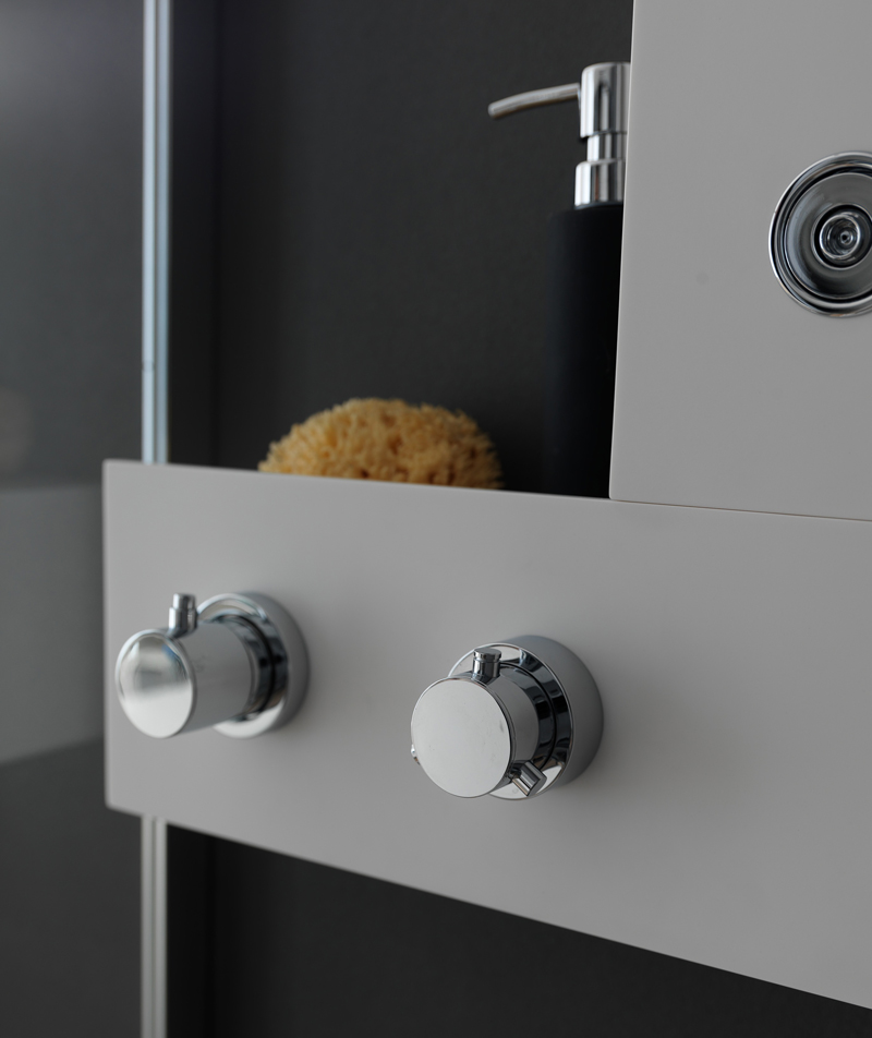 primo piano di miscelatore termostatico, deviatore e comandi cromati con modulo portaoggetti, su parete scura.