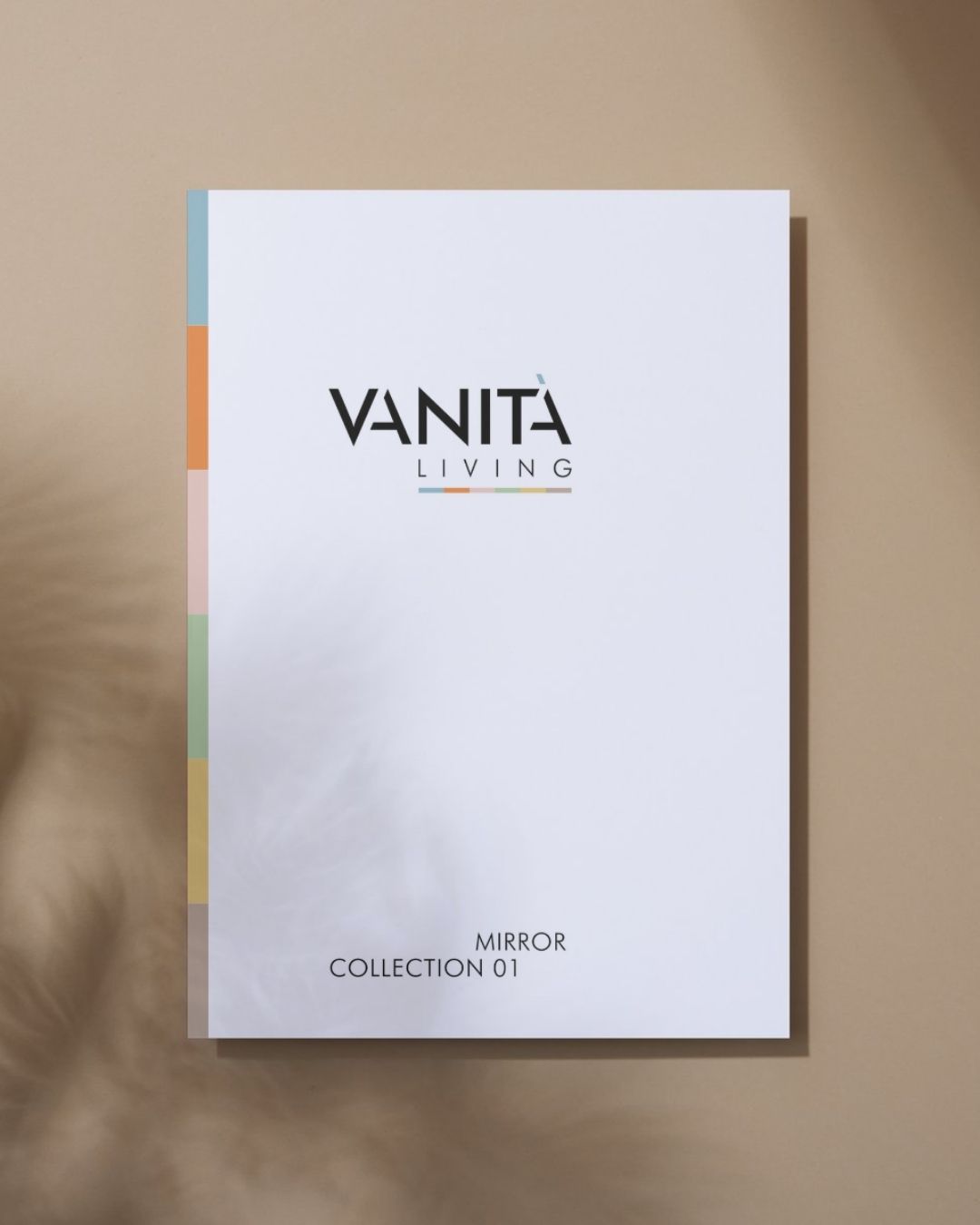 Copertina del catalogo Vanità Living appesa o appoggiata su fondo neutro beige, con logo centrale e striscia verticale di sei colori sul lato sinistro.