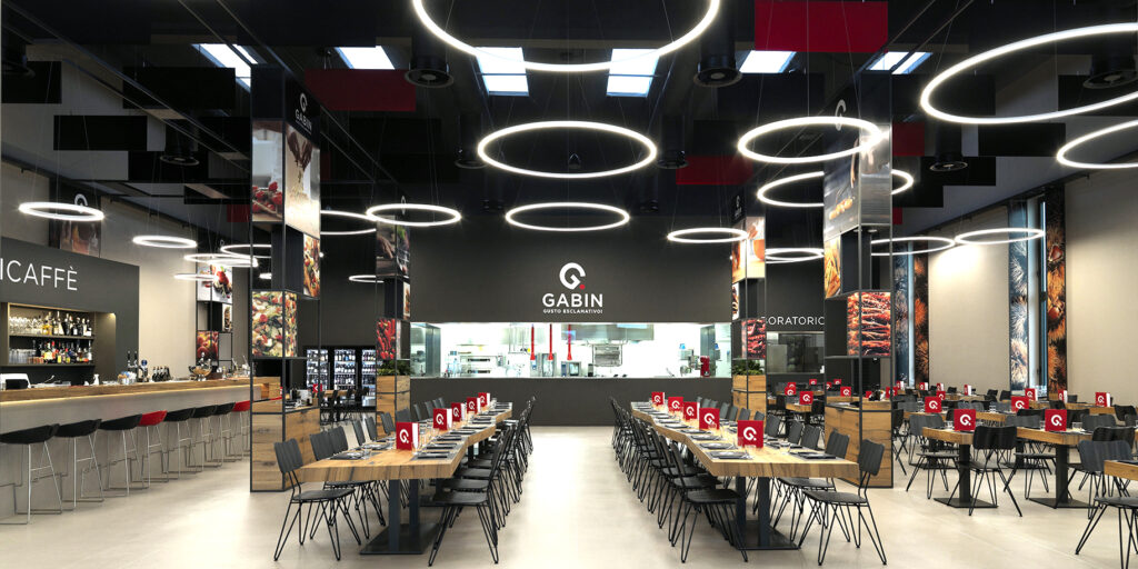 Vista interna ampia del ristorante Gabin Gusto Esclamativo! con tavoli, quinte metalliche e illuminazione sospesa in un ex capannone industriale.