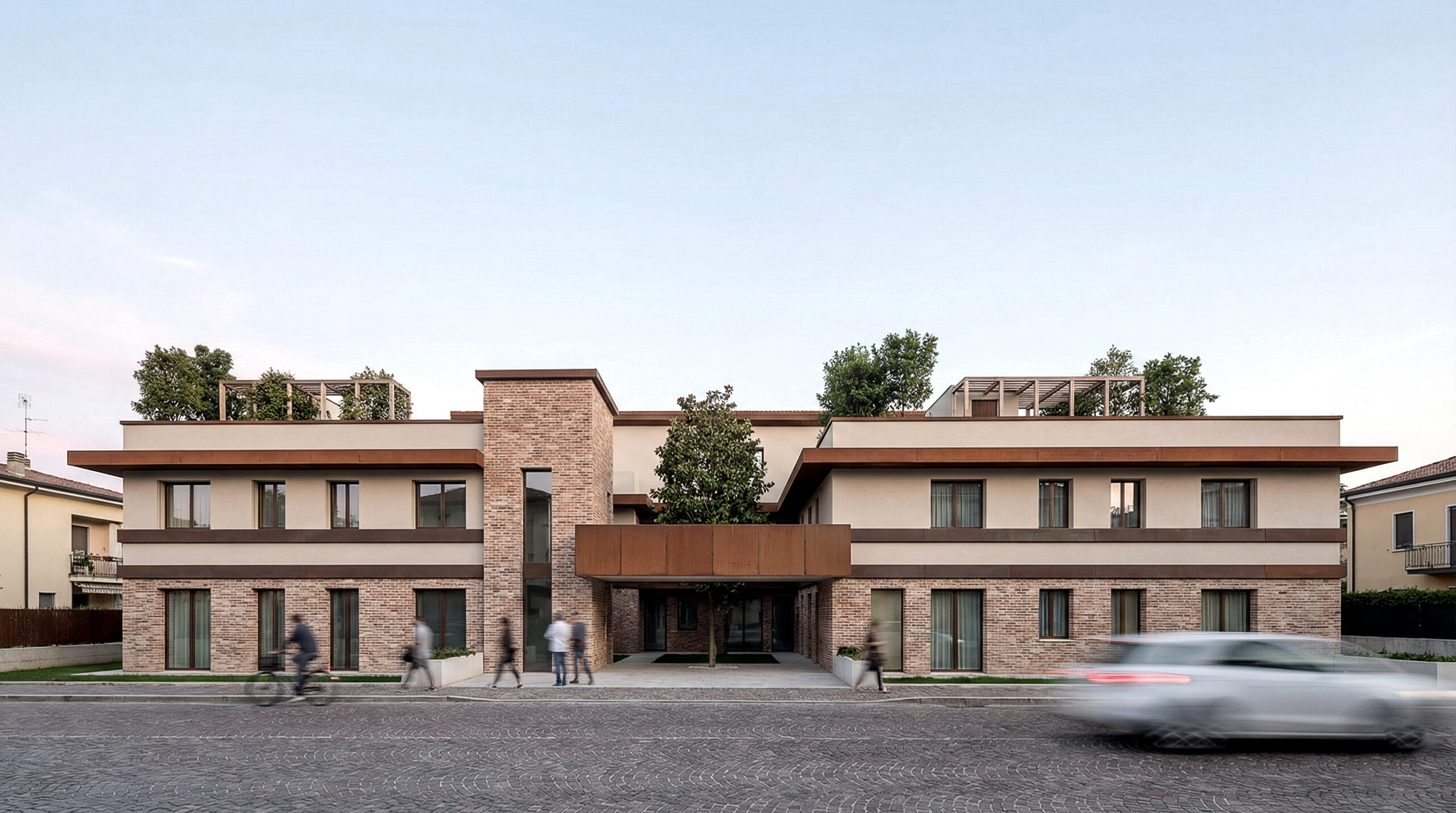 Vista frontale del progetto Housing Verona: edificio residenziale composto da due volumi orizzontali simmetrici in laterizio e intonaco chiaro, con ingresso centrale porticato, terrazze verdi in copertura, persone e auto in movimento sulla strada.