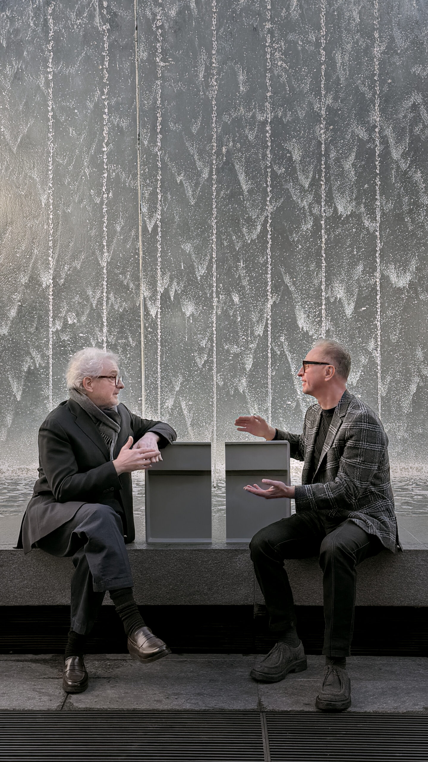 Il designer e Peteris Treicis, CEO dell’azienda, sono seduti davanti a una grande parete d’acqua mentre conversano; tra loro, appoggiati sul bordo in pietra, si vedono due prototipi grigi rettangolari della collezione Infinity.