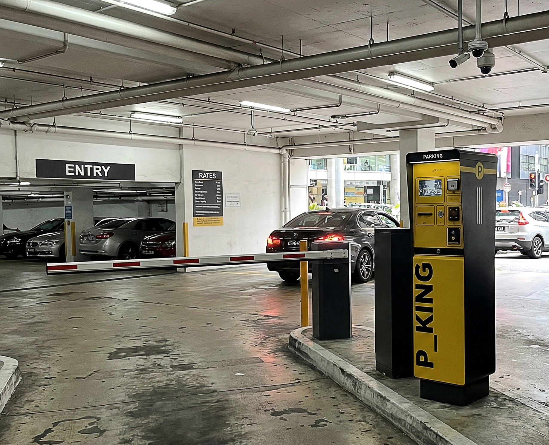 Vista ravvicinata di P_KING accanto a un’auto in ingresso a un parcheggio coperto, con conducente visibile nell’abitacolo, sbarra automatica e struttura in calcestruzzo sullo sfondo.