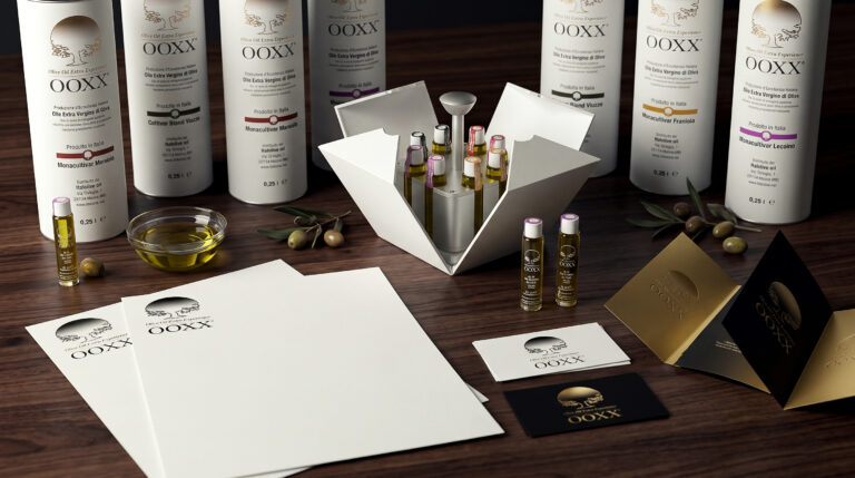 Branding OOXX Olive Oil Extra Experience: composizione hero con packaging cilindrico, oleobox aperta con flaconcini monodose in vetro, carta intestata, biglietti da visita e brochure su tavolo in legno con olio e olive.