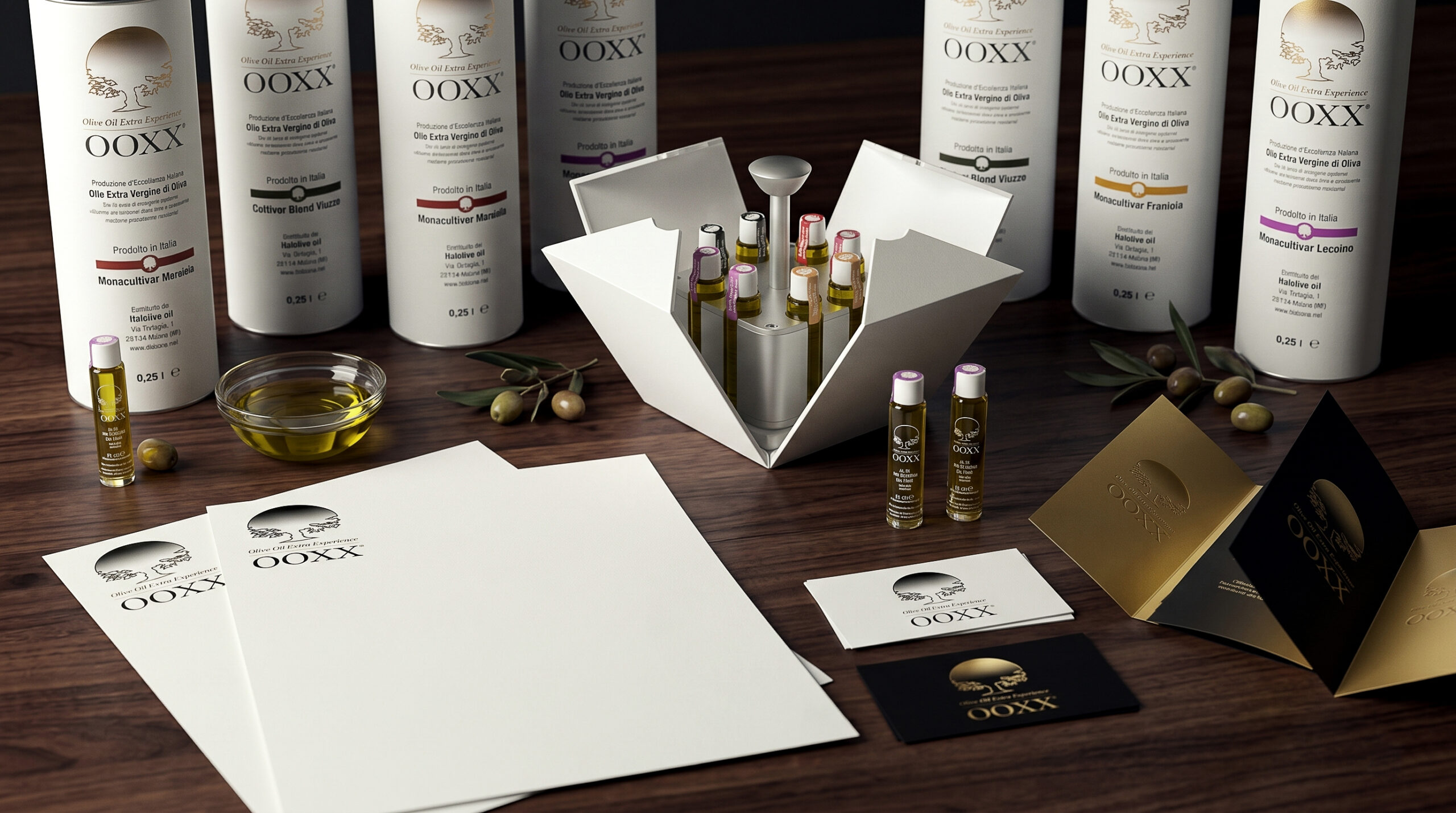 Branding OOXX Olive Oil Extra Experience: composizione hero con packaging cilindrico, oleobox aperta con flaconcini monodose in vetro, carta intestata, biglietti da visita e brochure su tavolo in legno con olio e olive.
